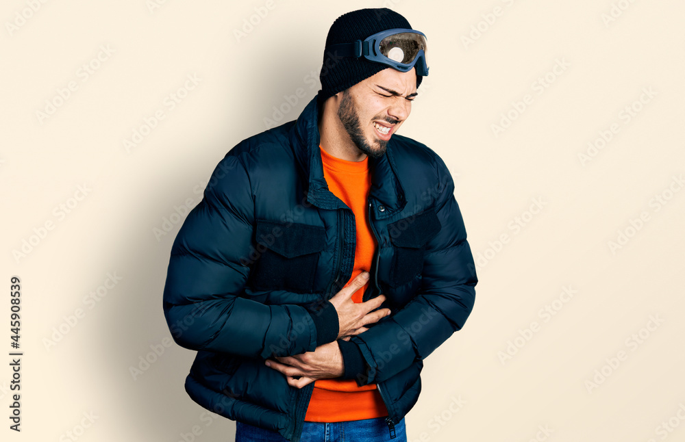 Young hispanic man with beard wearing snow wear and sky glasses with hand on stomach because nausea, painful disease feeling unwell. ache concept.