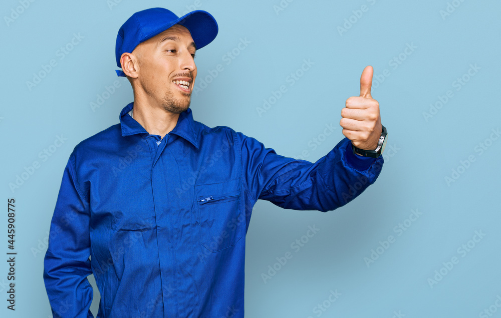 Bald man with beard wearing builder jumpsuit uniform looking proud, smiling doing thumbs up gesture to the side