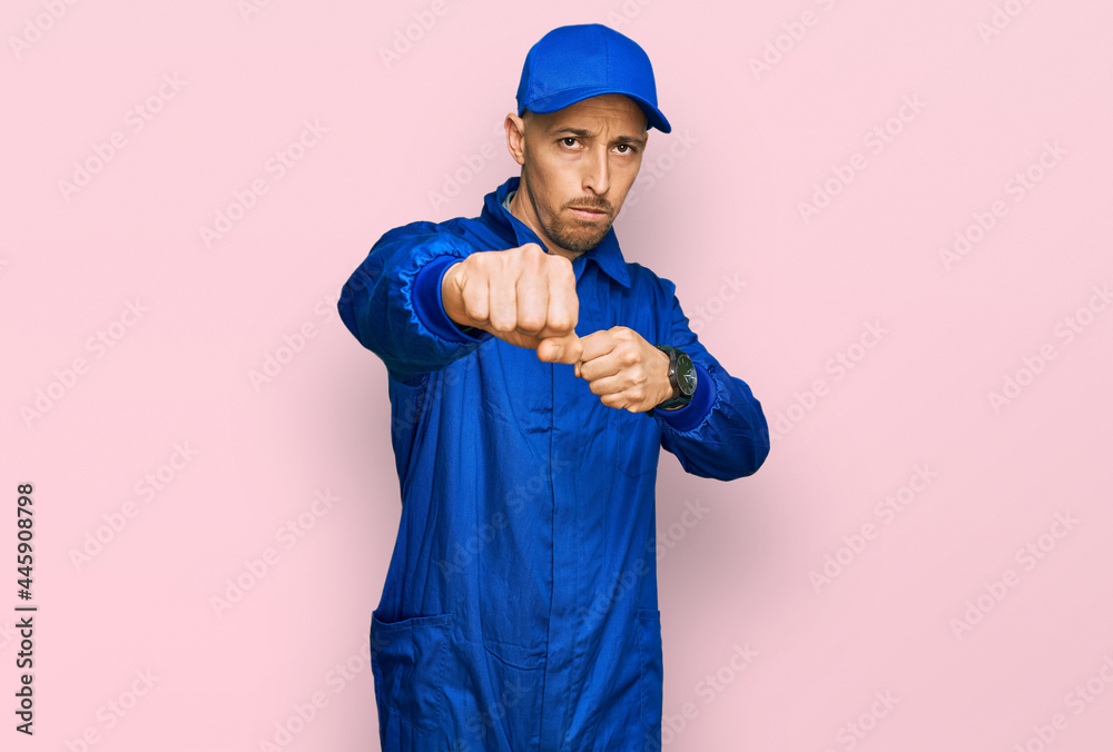Bald man with beard wearing builder jumpsuit uniform punching fist to fight, aggressive and angry attack, threat and violence