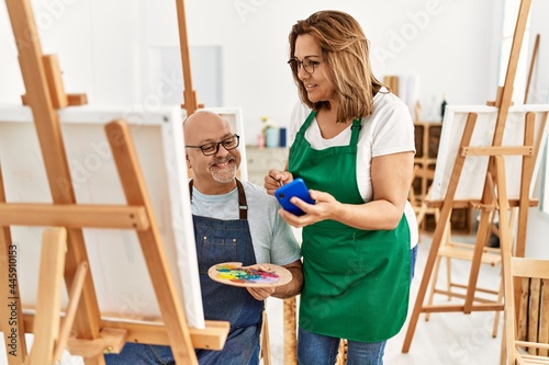 Middle age hispanic painter couple smiling happy painting and using smartphone at art studio.
