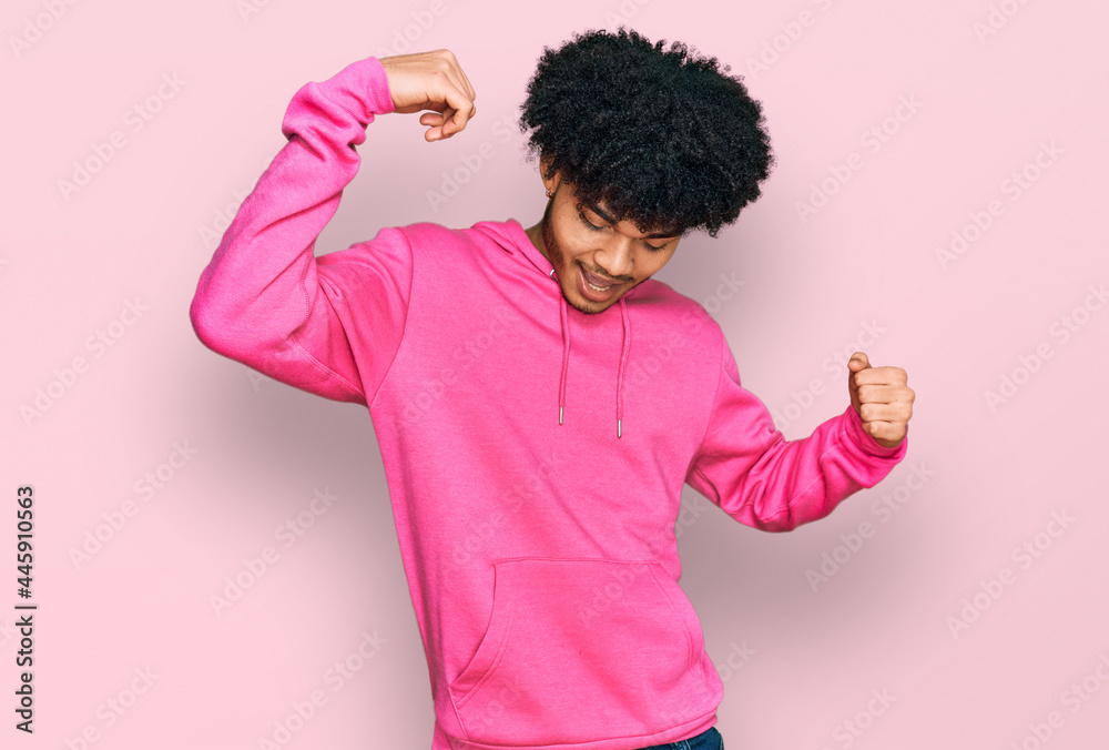 Young african american man with afro hair wearing casual pink sweatshirt dancing happy and cheerful, smiling moving casual and confident listening to music Young african american man with afro hair wearing casual pink sweatshirt dancing happy and cheerful, smiling moving casual and confident listening to music