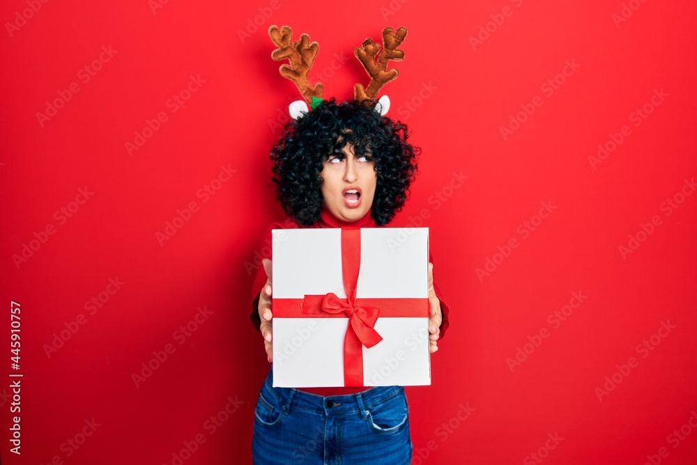 Young middle east woman wearing deer christmas hat holding gift angry and mad screaming frustrated and furious, shouting with anger. rage and aggressive concept.