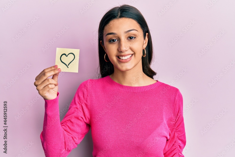 Young hispanic woman holding heart reminder looking positive and happy standing and smiling with a confident smile showing teeth Young hispanic woman holding heart reminder looking positive and happy standing and smiling with a confident smile showing teeth