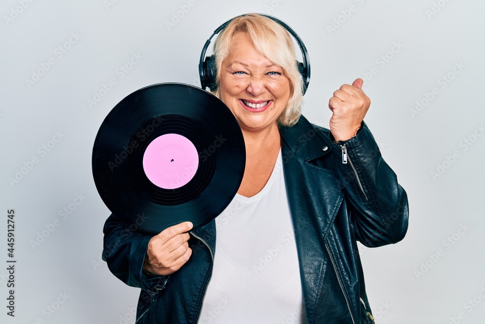 Middle age blonde woman holding vinyl disc using headphones screaming proud, celebrating victory and success very excited with raised arm Middle age blonde woman holding vinyl disc using headphones screaming proud, celebrating victory and success very excited with raised arm