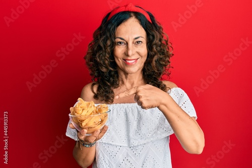 Middle age hispanic woman holding bowl with uncooked pasta smiling happy pointing with hand and finger