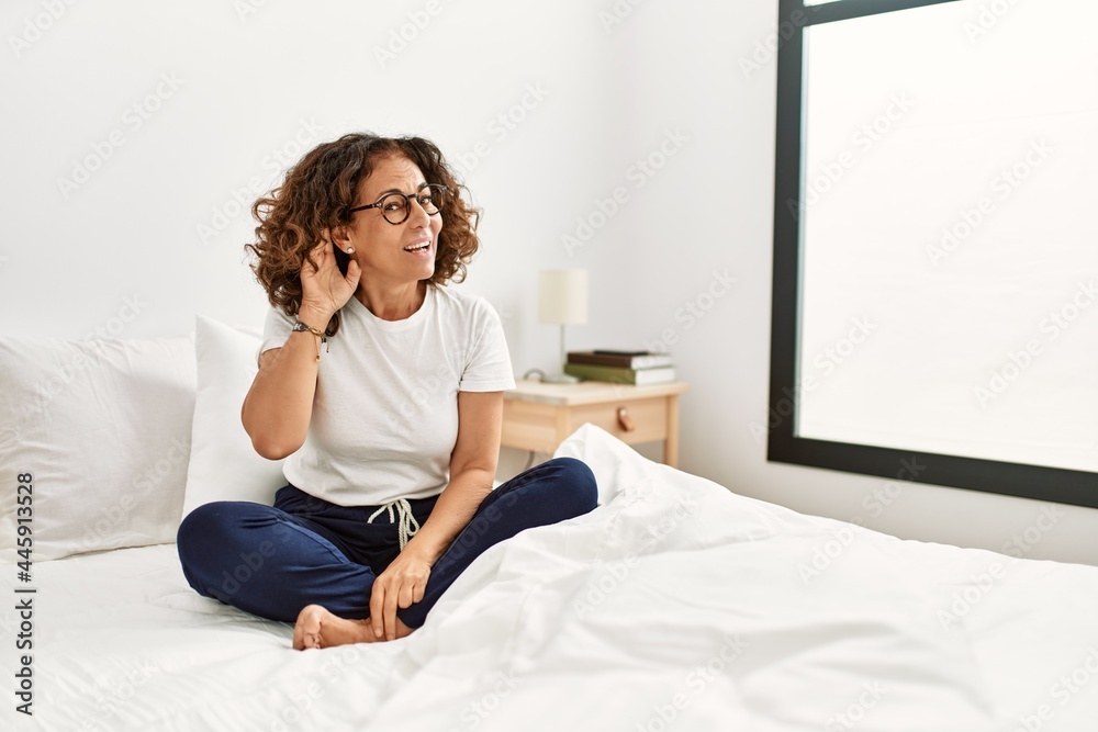Middle age hispanic woman sitting on the bed at home smiling with hand over ear listening an hearing to rumor or gossip. deafness concept.