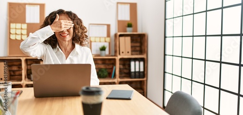 Middle age hispanic woman working at the office wearing glasses smiling and laughing with hand on face covering eyes for surprise. blind concept.