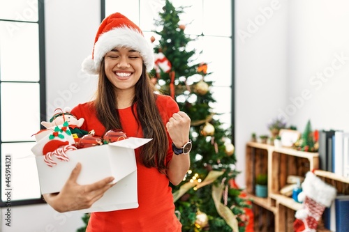Young brunette woman standing by christmas tree holding decoration very happy and excited doing winner gesture with arms raised  smiling and screaming for success. celebration concept.