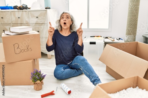 Middle age woman with grey hair sitting on the floor at new home amazed and surprised looking up and pointing with fingers and raised arms. photo