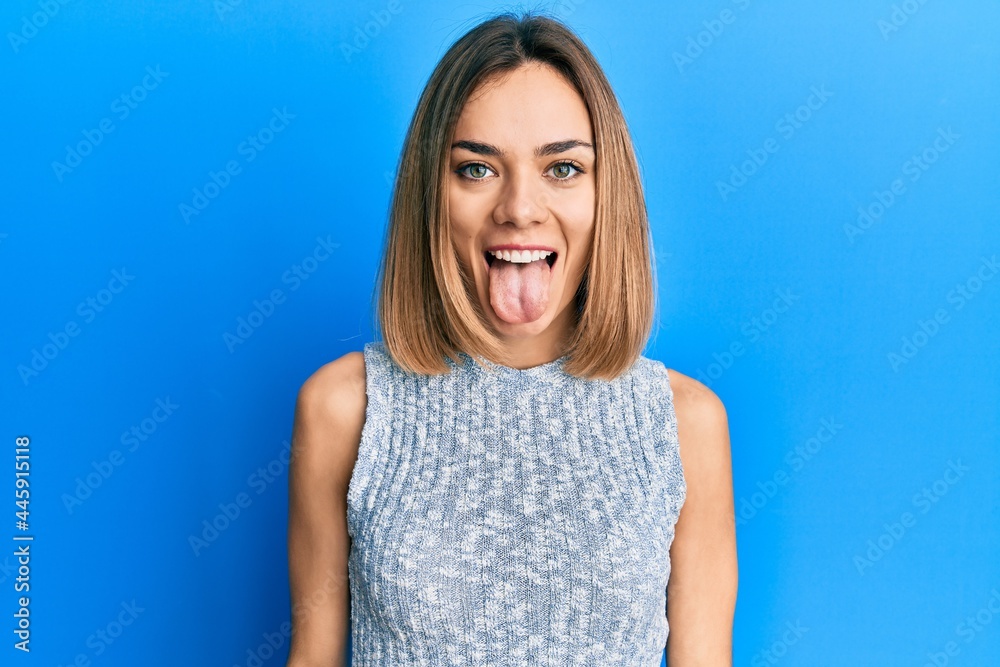 Young caucasian blonde woman wearing casual t shirt sticking tongue out happy with funny expression. emotion concept. Young caucasian blonde woman wearing casual t shirt sticking tongue out happy with funny expression. emotion concept.