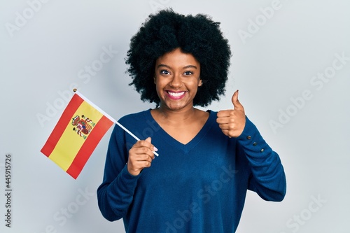Young african american woman holding spain flag smiling happy and positive, thumb up doing excellent and approval sign
