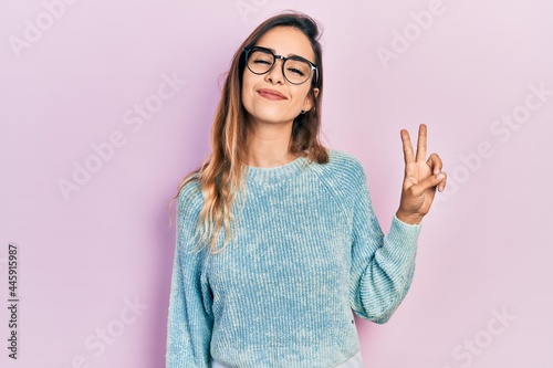 Young hispanic girl wearing casual clothes and glasses smiling with happy face winking at the camera doing victory sign. number two.