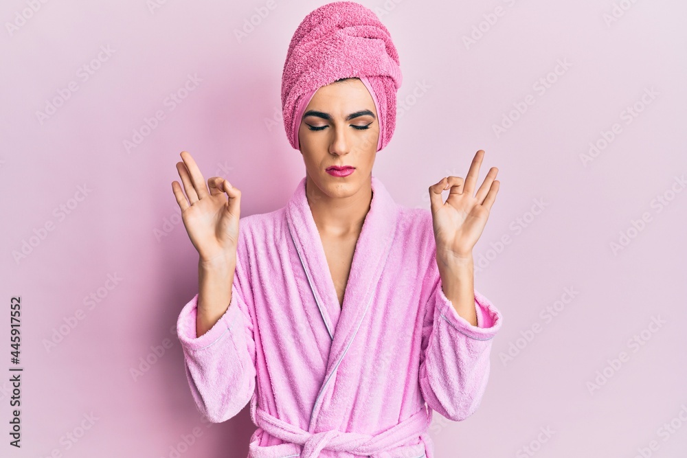 Young man wearing woman make up wearing shower towel on head and bathrobe relaxed and smiling with eyes closed doing meditation gesture with fingers. yoga concept. Young man wearing woman make up wearing shower towel on head and bathrobe relaxed and smiling with eyes closed doing meditation gesture with fingers. yoga concept.