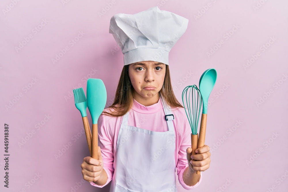 Fototapeta premium Beautiful brunette little girl wearing professional cook apron holding cooking tools depressed and worry for distress, crying angry and afraid. sad expression.
