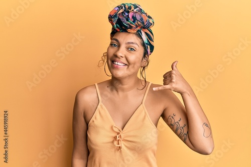 Young hispanic girl wearing hair turban over yellow background smiling doing phone gesture with hand and fingers like talking on the telephone. communicating concepts.