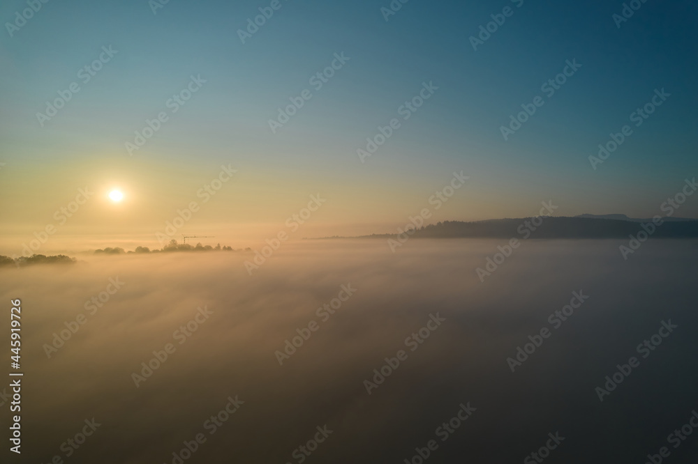 Landscape shot above the fog. Yellow sun rising on horizon. Panorama with trees and hills. A construction crane is visible. Cyan colors. Aerial view.