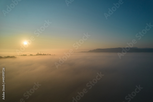 Landscape shot above the fog. Yellow sun rising on horizon. Panorama with trees and hills. A construction crane is visible. Cyan colors. Aerial view. © Jan