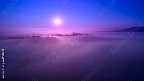 The sun rises above the fog. Small village surrounded by trees. Landscape fantasy designed with blue colors. Aerial view.