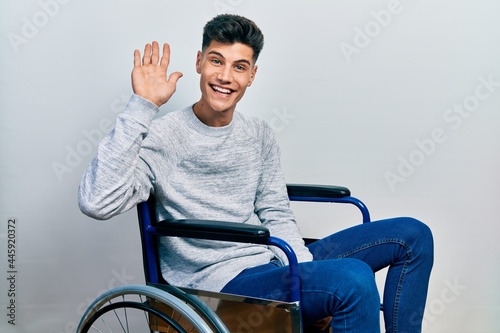 Young hispanic man sitting on wheelchair waiving saying hello happy and smiling, friendly welcome gesture