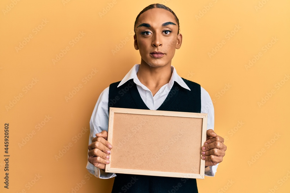 Hispanic man wearing make up and long hair holding empty corkboard relaxed with serious expression on face. simple and natural looking at the camera.