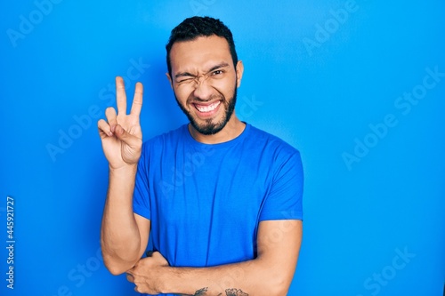 Hispanic man with beard wearing casual blue t shirt smiling with happy face winking at the camera doing victory sign. number two.