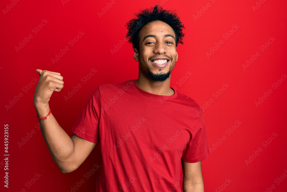 Young african american man with beard wearing casual red t shirt smiling with happy face looking and pointing to the side with thumb up. Young african american man with beard wearing casual red t shirt smiling with happy face looking and pointing to the side with thumb up.