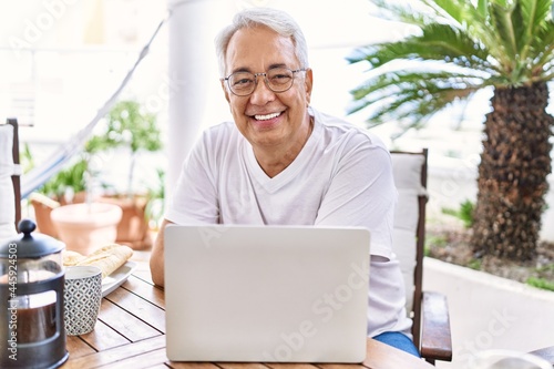 Handsome middle age hispanic man with grey hair and glasses working using computer laptop at home. Smiling happy and confident, relaxed at house terrace.