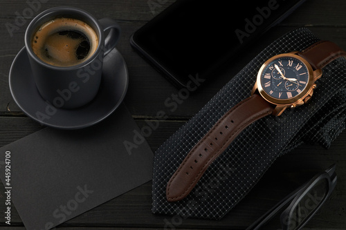men's wrist watch on a black tie, a hot cup of coffee and a smartphone on a wooden table. black envelope with place for text.