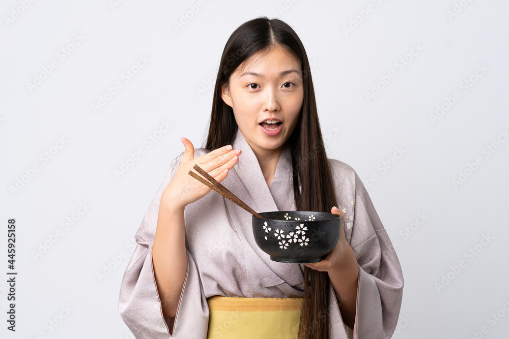 Young Chinese girl wearing kimono over isolated white background holding a bowl of noodles with chopsticks and surprised Young Chinese girl wearing kimono over isolated white background holding a bowl of noodles with chopsticks and surprised