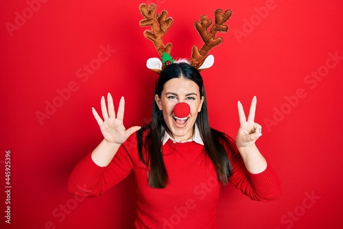 Young hispanic woman wearing deer christmas hat and red nose showing and pointing up with fingers number seven while smiling confident and happy.