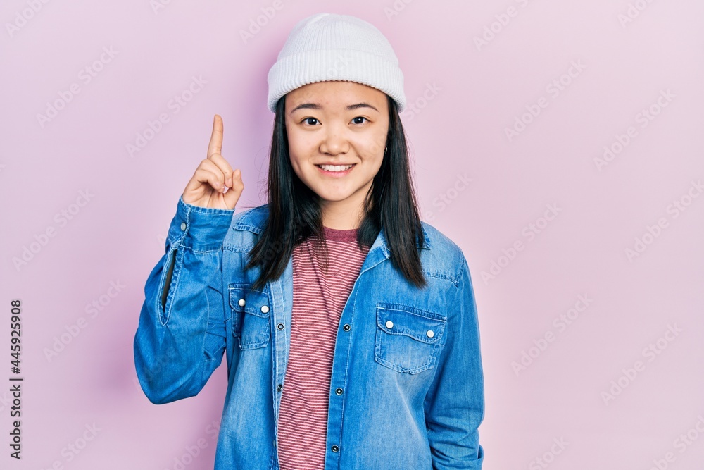 Young chinese girl wearing cute wool cap pointing finger up with successful idea. exited and happy. number one. Young chinese girl wearing cute wool cap pointing finger up with successful idea. exited and happy. number one.