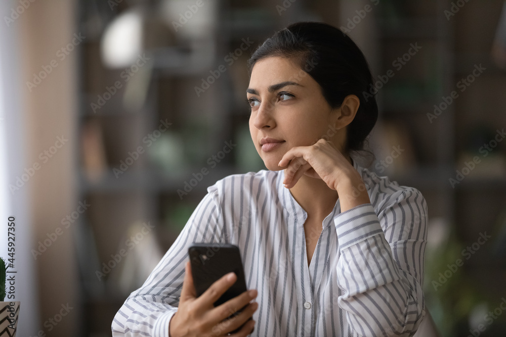 Pensive young Indian woman use cellphone gadget look in distance thinking or planning. Thoughtful millennial mixed race female hold smartphone text message visualizing. Technology concept. Pensive young Indian woman use cellphone gadget look in distance thinking or planning. Thoughtful millennial mixed race female hold smartphone text message visualizing. Technology concept.