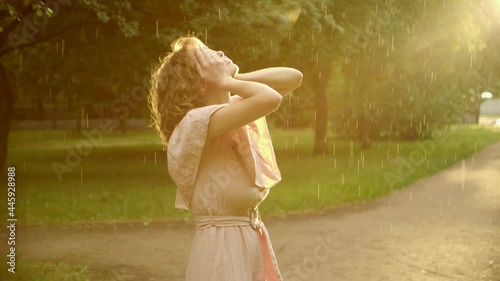 a woman in a dress, enjoys the rain in the light of the departing sun, she raises her hands and covers her face with happiness, the image of a beautiful youth, youth of the soul