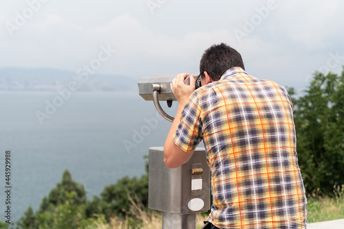 young man using stationary binoculars on a warm summer day on a hill against the background of the mountains and sea