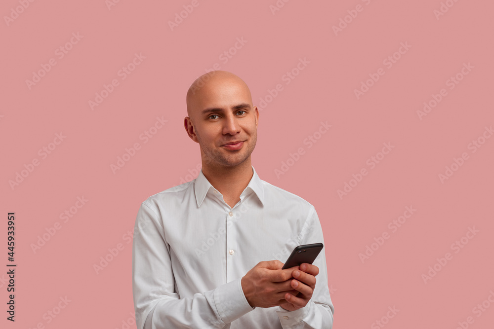 Good looking bald man possing over pink studio background, holds smartphone, looking at the camera. Likes to communicate with friends by phone. Has bristle. Dressed in white shirt. Good looking bald man possing over pink studio background, holds smartphone, looking at the camera. Likes to communicate with friends by phone. Has bristle. Dressed in white shirt.