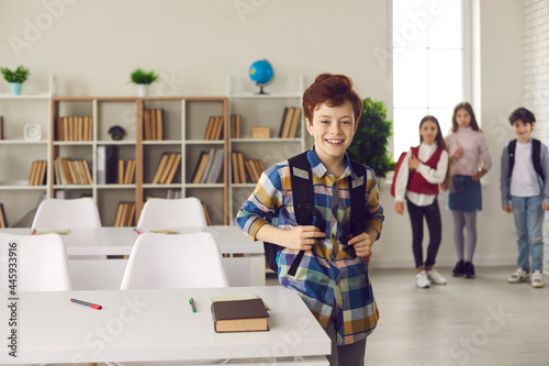 Elementary students back to school. Smiling caucasian schoolboy with backpack standing next to desk posing for camera at bright classroom. Diverse classmates waiting on blurred background