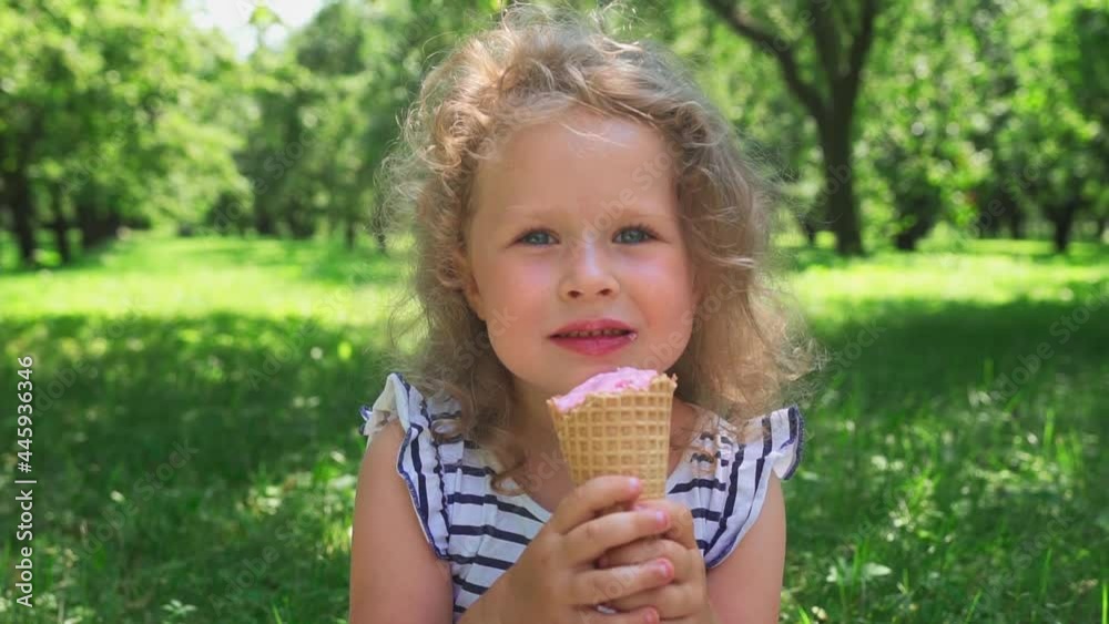 Cute blonde curly girl eating ice cream in the park on a sunny summer day. A child enjoys ice cream in a waffle cone during the heatwave outdoors. Slow motion.