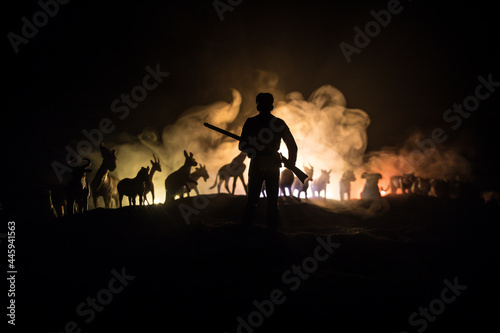Silhouette of a man (hunter) with rifle standing against group of animals in colorful dark backlight. Decorated with miniatures. Selective focus
