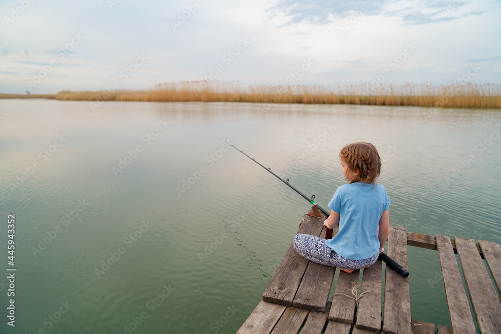 rear view. kid girl sits on a wooden bridge on a river with fishing rod. rear view. kid girl sits on a wooden bridge on a river with fishing rod.