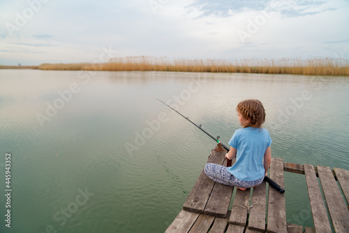 rear view. kid girl sits on a wooden bridge on a river with fishing rod.