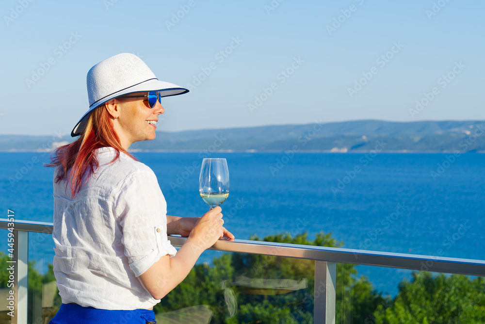 beautiful woman in straw hat enjoying evening with wine glass at resort. Luxury hotel terrace. Europe summer vacation. Happy woman drinking white wine relaxing enjoying view of the mediterranean blue
