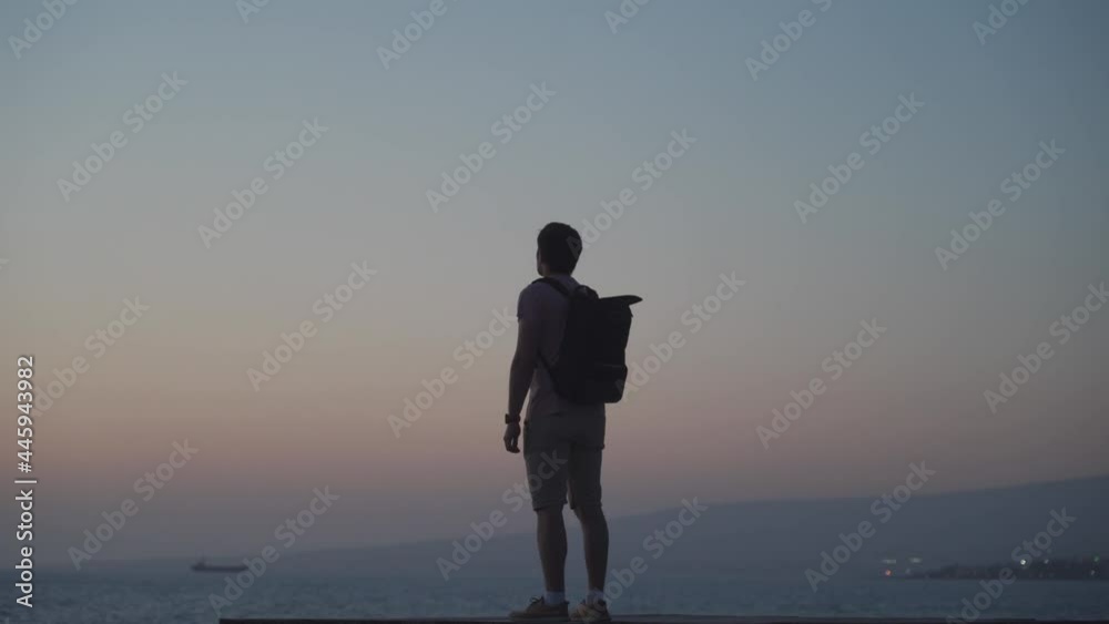 Theme of loneliness, depression, search for answers to questions while traveling. Man traveler with backpack looks at sunset of Mediterranean sea from wooden pier on island of Cyprus, city of Paphos.
