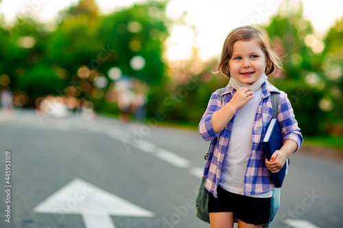 a schoolgirl is standing on the road in a plaid shirt and a medical mask, with a school backpack and a diary in her hands