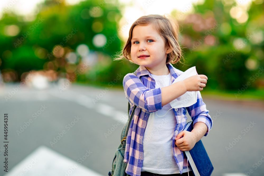 a schoolgirl is standing on the road in a plaid shirt and a medical mask, with a school backpack and a diary in her hands a schoolgirl is standing on the road in a plaid shirt and a medical mask, with a school backpack and a diary in her hands