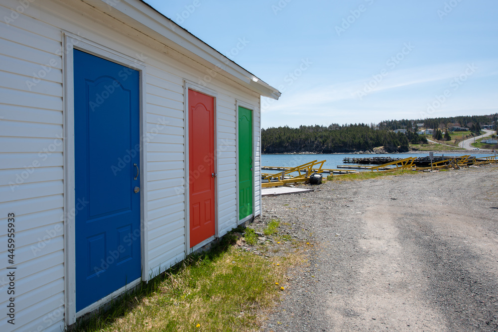 A row of colorfully painted solid doors of blue, red, and green. The exterior wall is white vinyl siding. The sky is blue in the background and the storage units are sitting on gravel with blue ocean.
