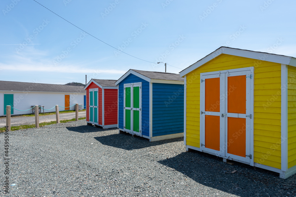 A row of small colorful painted huts or sheds made of wood. The exterior walls are colorful with double wooden doors. The sky is blue in the background and the storage units are sitting on gravel.