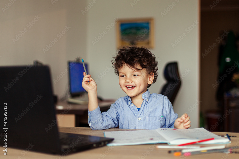 a happy preschool child is trained online on a laptop and does homework in a notebook. A cute, joyful curly boy looks at the monitor screen in a room of his house a happy preschool child is trained online on a laptop and does homework in a notebook. A cute, joyful curly boy looks at the monitor screen in a room of his house