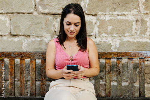 Young woman using mobile phone while sitting on bench at park - Cheerful hispanic student texting and reading message on her smartphone outdoor - Social media concept
