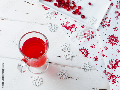 Selective focus on cranberries in a fresh drink in a glass cup. Berries on a white rectangular ceramic plate. Napkin with Christmas ornament. New Year's decor on a white wooden background. Copy space.
