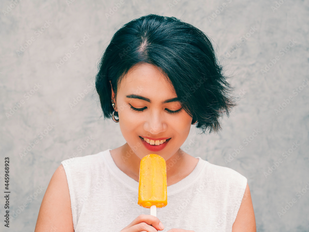 Smiling Asian woman short hair in casual white sleeveless shirt holding yellow popsicle on grey concrete wall background in summertime. Women eating popsicles, freshy summer season concept. Smiling Asian woman short hair in casual white sleeveless shirt holding yellow popsicle on grey concrete wall background in summertime. Women eating popsicles, freshy summer season concept.